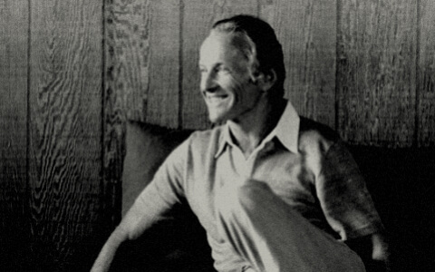 A black and white photograph of a smiling man wearing a collared shirt. He sits in front of a wood-paneled wall.