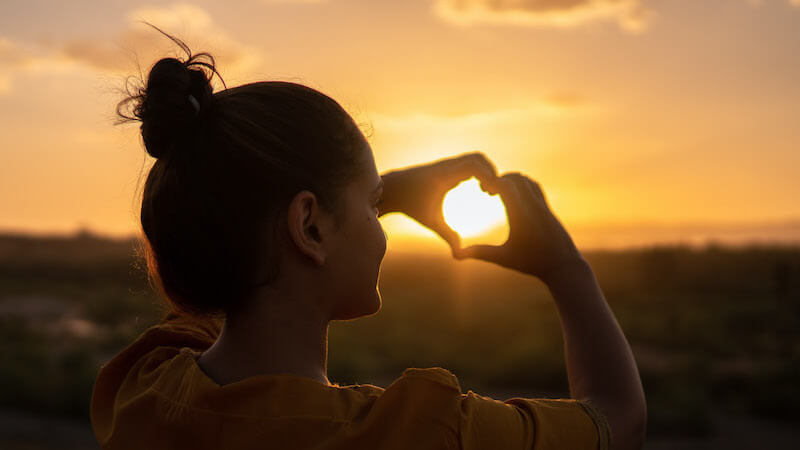 Photo of a person outdoors during sunset, holding their hands in a heart shape.